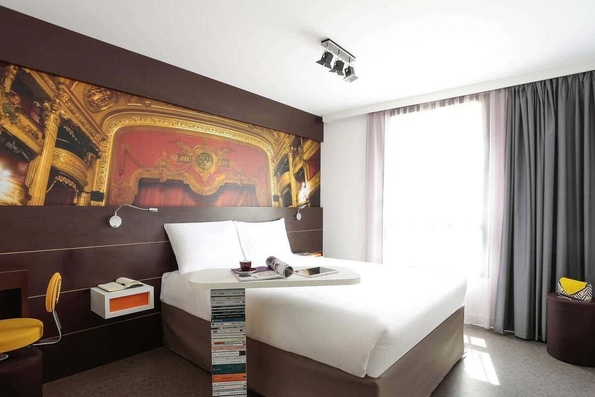 Ein Hotelzimmer mit einem großen Bett, einem Kopfteil mit Wandgemäldeverzierung, einem Nachttisch mit Lampe und einem Fenster mit Vorhängen.