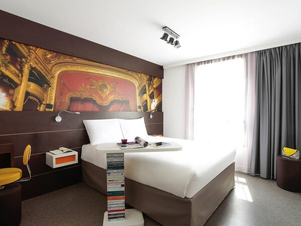 Una habitación de hotel con una cama grande, un cabecero con un mural decorativo, una mesita de noche con una lámpara y una ventana con cortinas.
