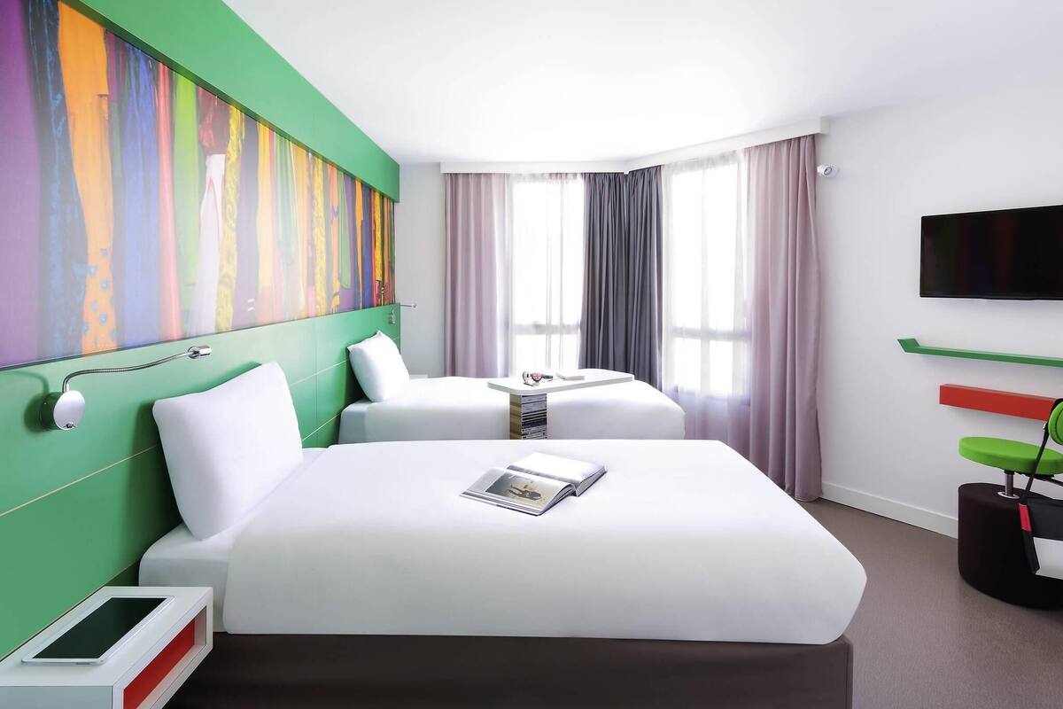 Ein Hotelzimmer mit einem großen Bett, einem Nachttisch mit einem Tablet, einem an der Wand montierten Fernseher und einem farbenfrohen abstrakten Gemälde.