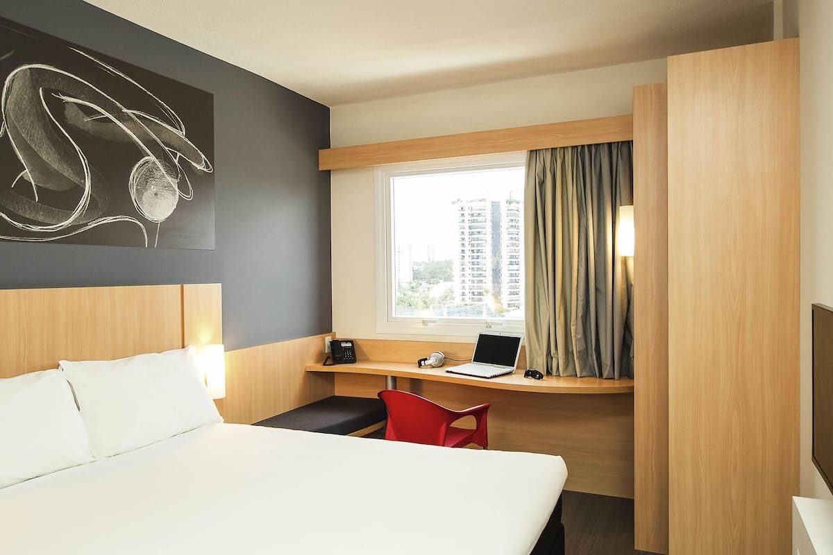 Ein Hotelzimmer mit einem Bett, einem Schreibtisch mit einem Laptop, einem Sessel, einem Telefon und einem Fenster mit Vorhängen.