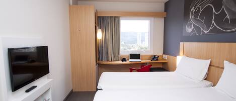 A hotel room with a TV, a desk with a laptop, a bed, and a window with curtains.