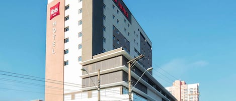 A modern hotel building with a prominent 'ibis Hotel' sign, multiple floors, and a flat roof design.