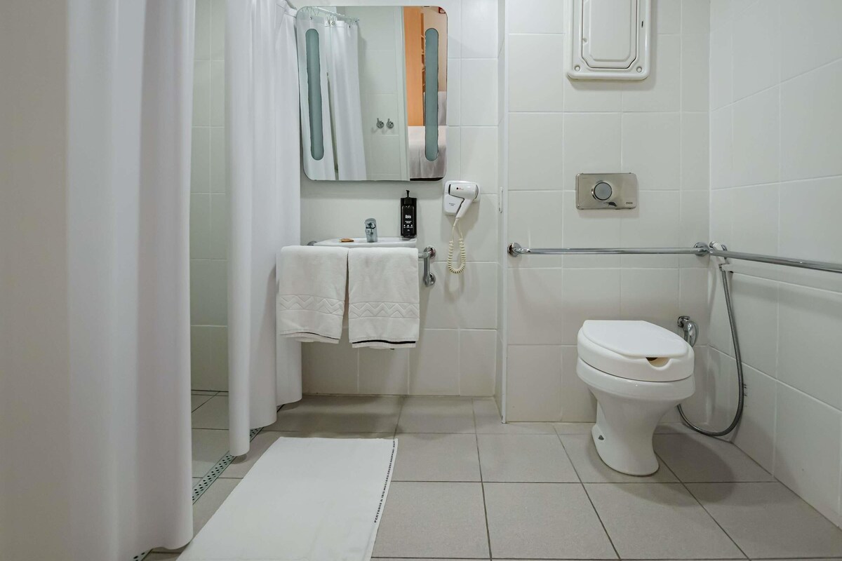 Un baño con inodoro, lavamanos con toalla, cortina de ducha, espejo y cabezal de ducha de mano.