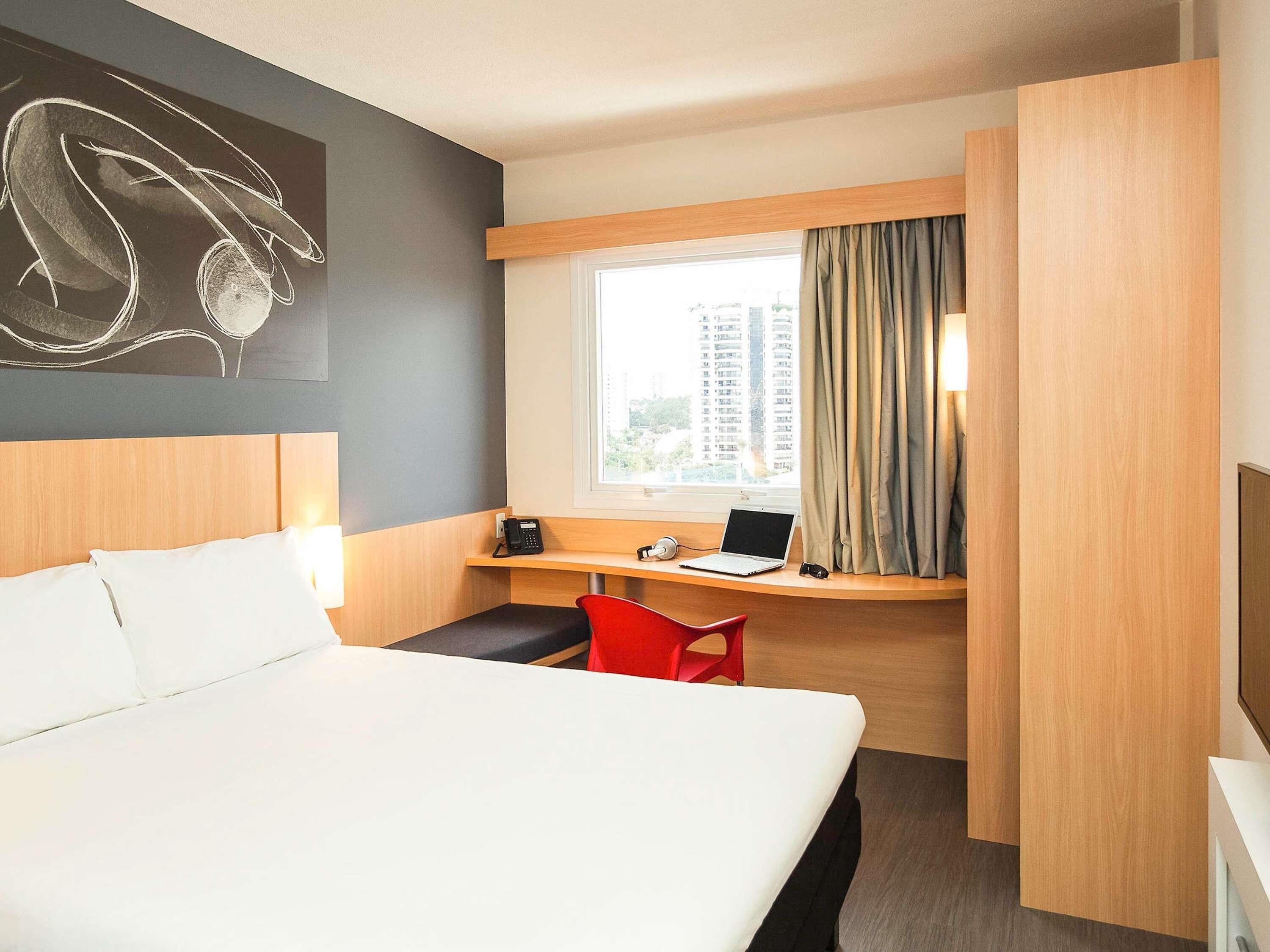 Ein Hotelzimmer mit einem Bett, einem Schreibtisch mit einem Laptop, einem Telefon und einem roten Sessel.