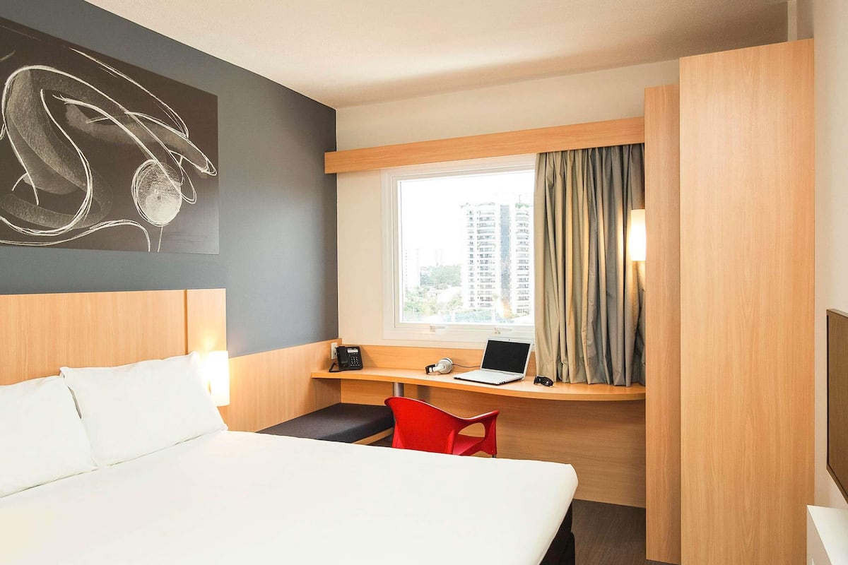 A hotel room with a bed, a desk with a laptop, a telephone, and a red chair.