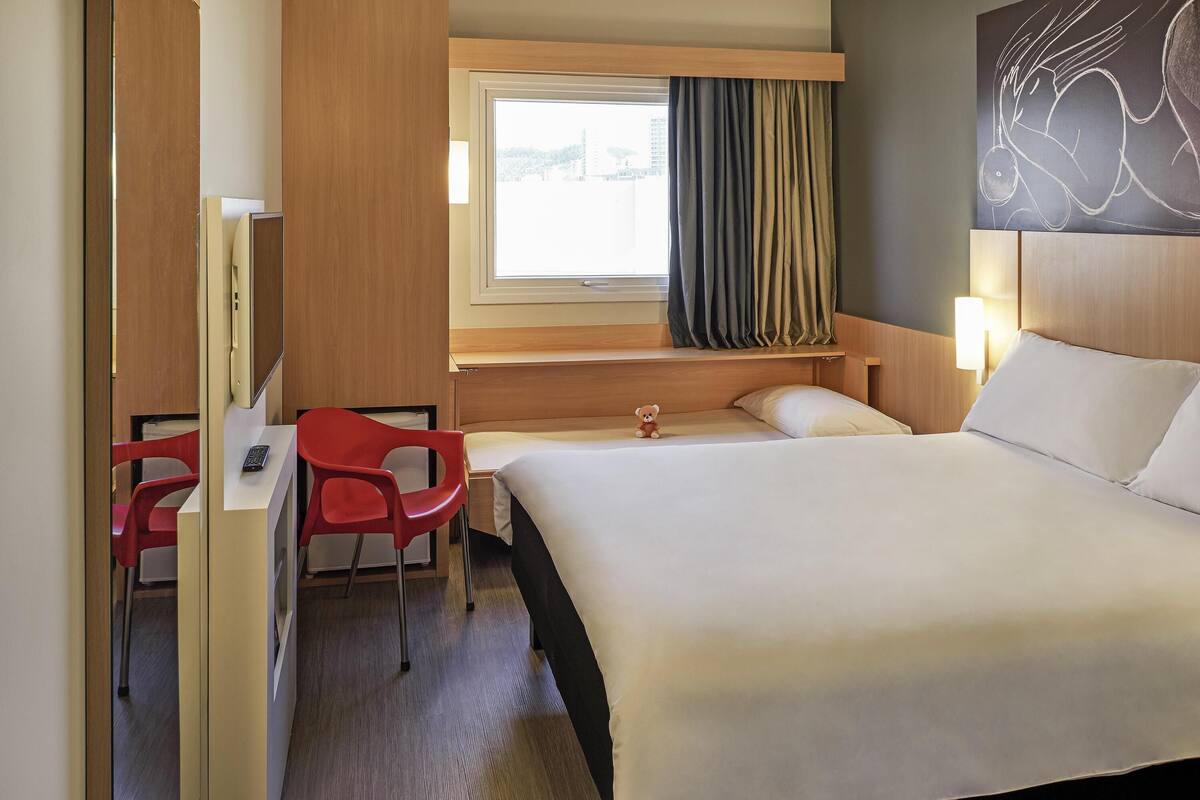 Une chambre d’hôtel comprenant un lit, un bureau avec une chaise, une fenêtre avec des rideaux et une œuvre d’art accrochée au mur.