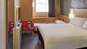 A hotel room with a bed, a desk with a chair, a window with curtains, and a wall-mounted artwork.