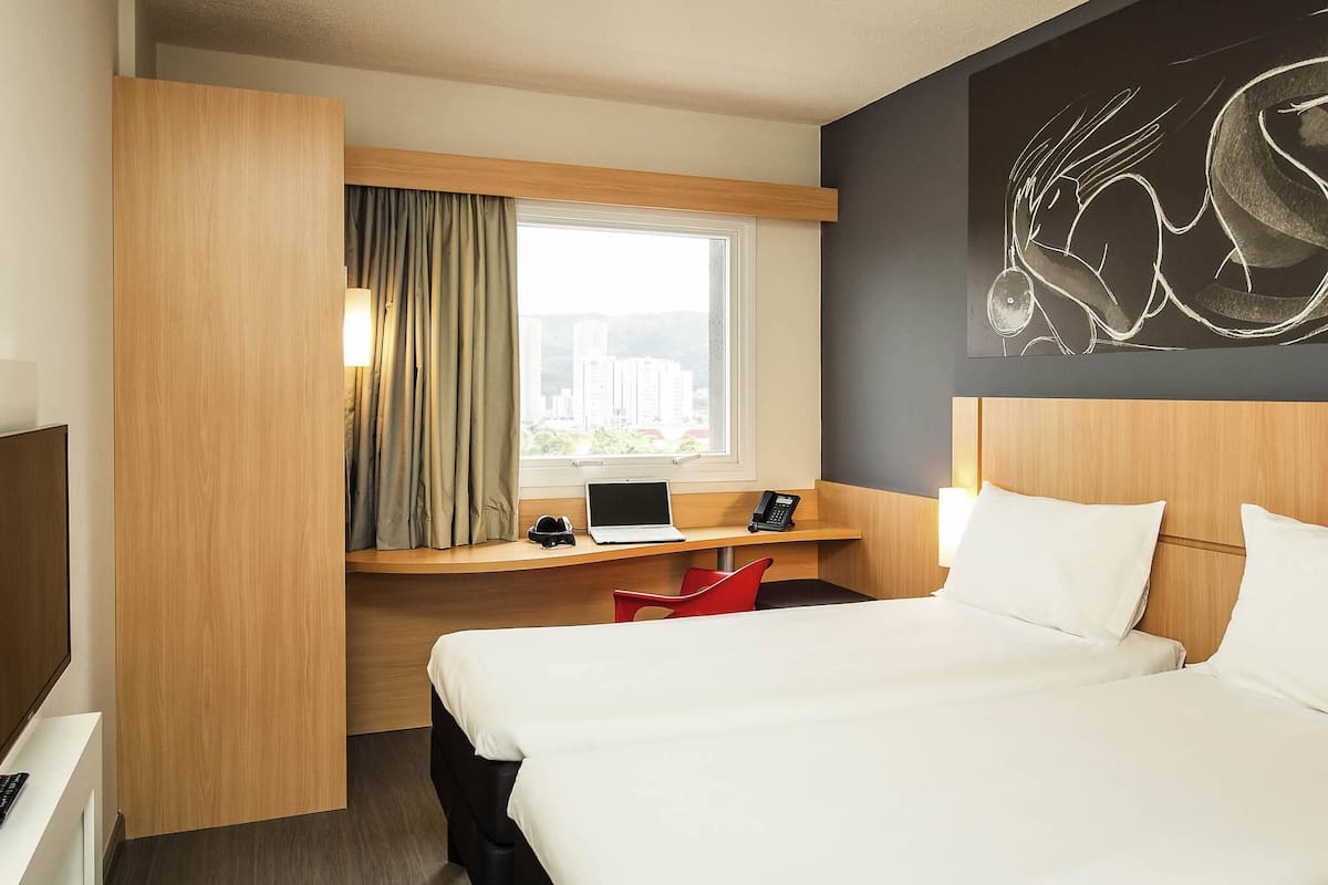 Ein Hotelzimmer mit einem Bett, einem Schreibtisch mit Laptop, einem Stuhl und einem Fenster mit Vorhängen.