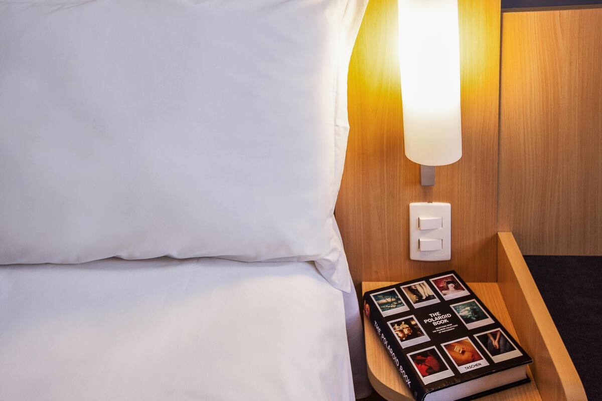 A hotel room with a neatly made bed, a bedside lamp, and a Polaroid book on a wooden nightstand.