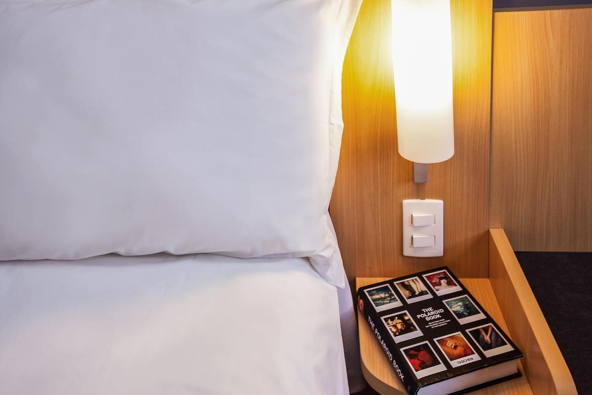 A hotel room with a neatly made bed, a bedside lamp, and a Polaroid book on a wooden nightstand.