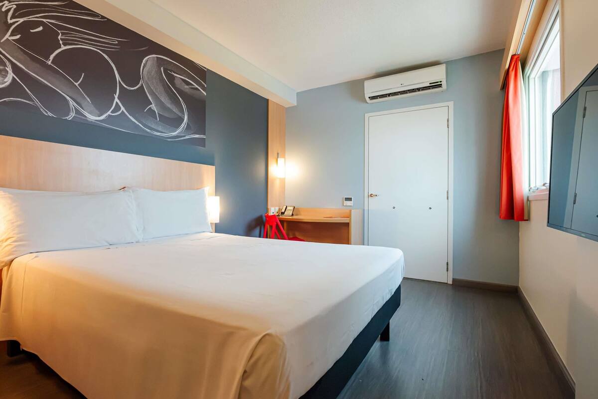 A hotel room with a bed, a desk, an air conditioning unit, a wardrobe, and a wall with a graphic.