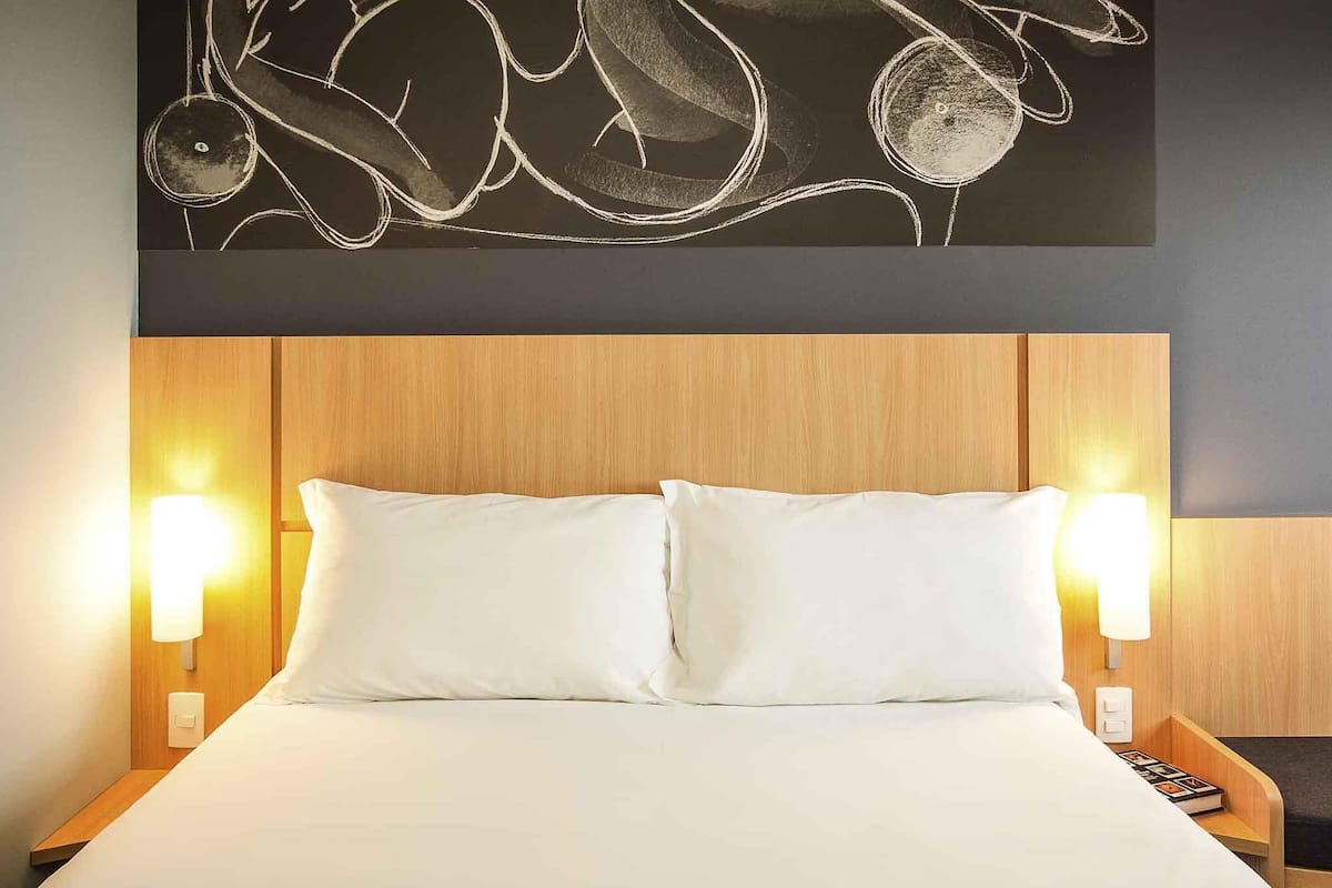 A hotel room with a bed, two bedside lamps, a wooden headboard, and a wall art piece.