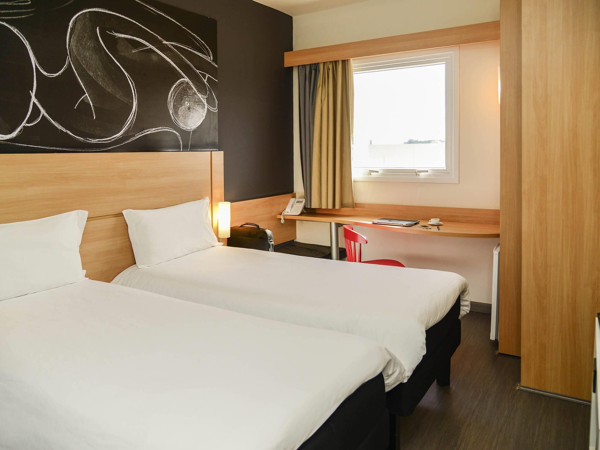 A hotel room with two beds, a desk, a chair, a window with curtains, and a wall art piece.