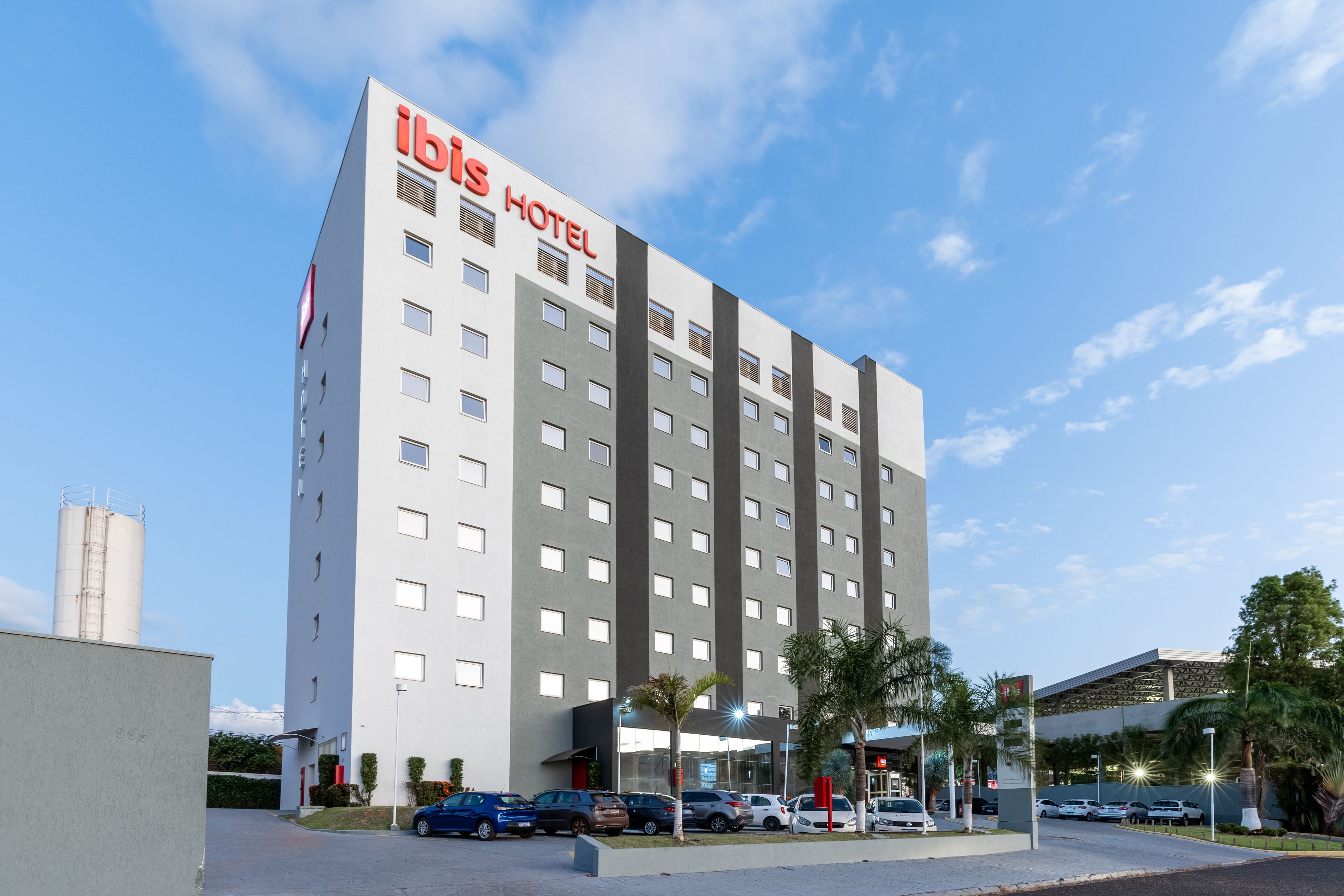 A modern hotel with a prominent 'ibis HOTEL' sign, multiple floors, and a parking area with several cars.
