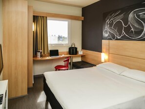 Una habitación de hotel con una cama, un escritorio con una laptop, una silla roja, una ventana con cortinas y una pared con una obra de arte abstracta de gran tamaño.