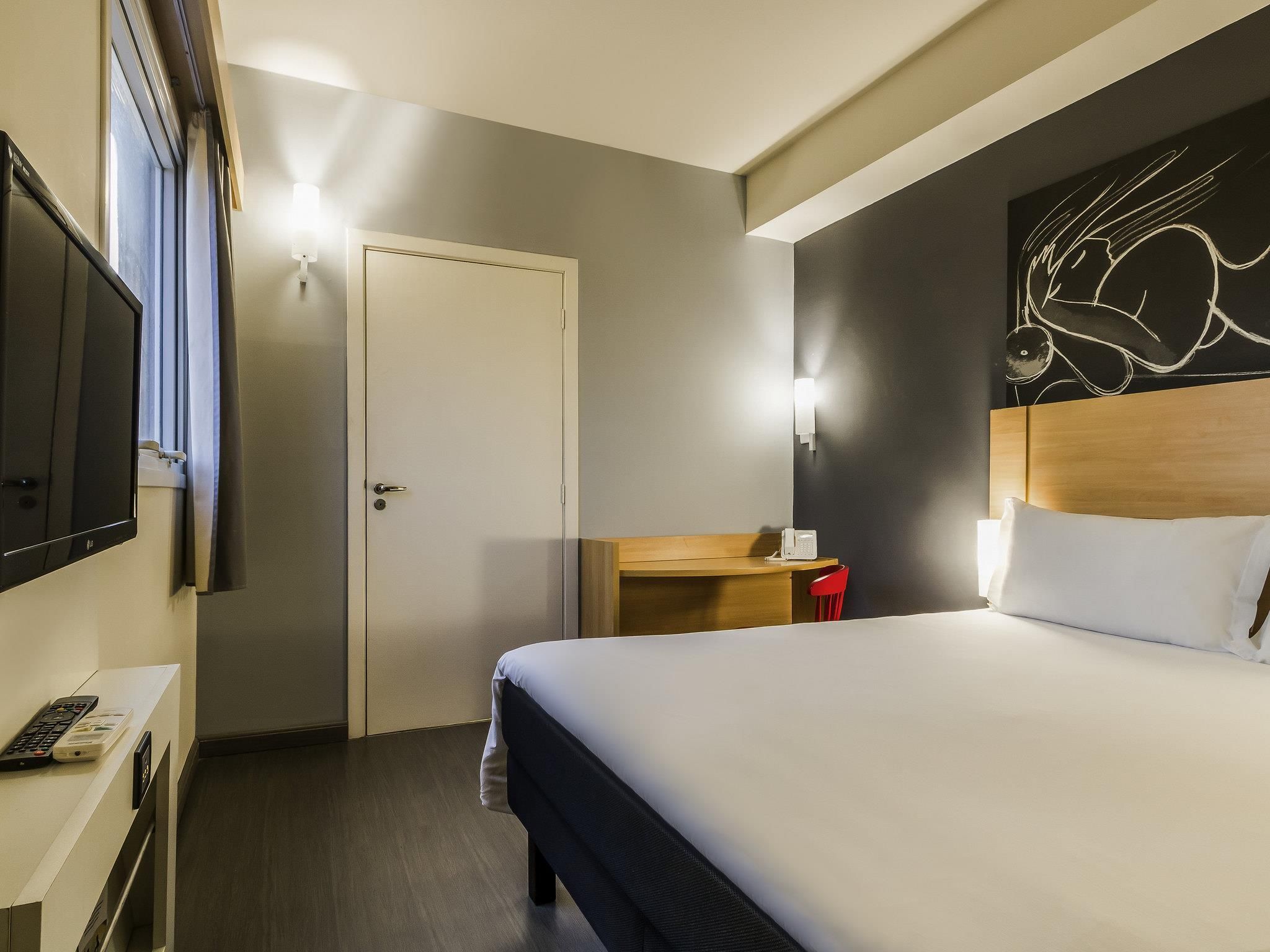 Quarto de hotel com cama, escrivaninha, cadeira, televisão e porta.