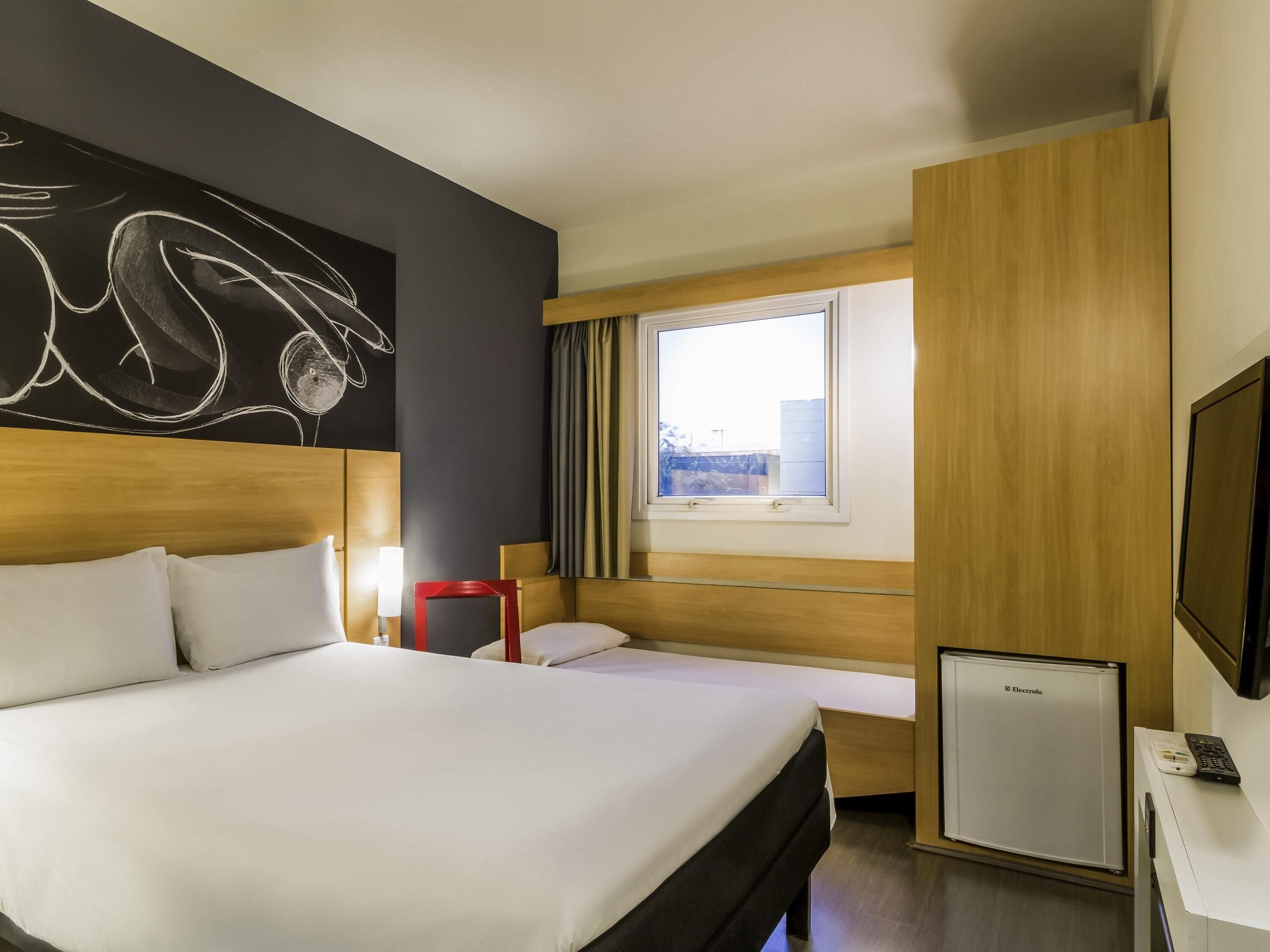 A hotel room with a large bed, a bedside table with a lamp, a small desk with a refrigerator and a TV, and a window with curtains.