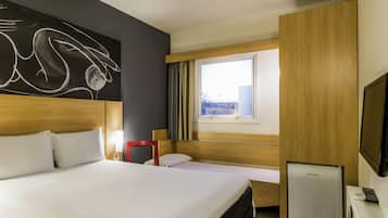 A hotel room with a large bed, a bedside table with a lamp, a small desk with a refrigerator and a TV, and a window with curtains.