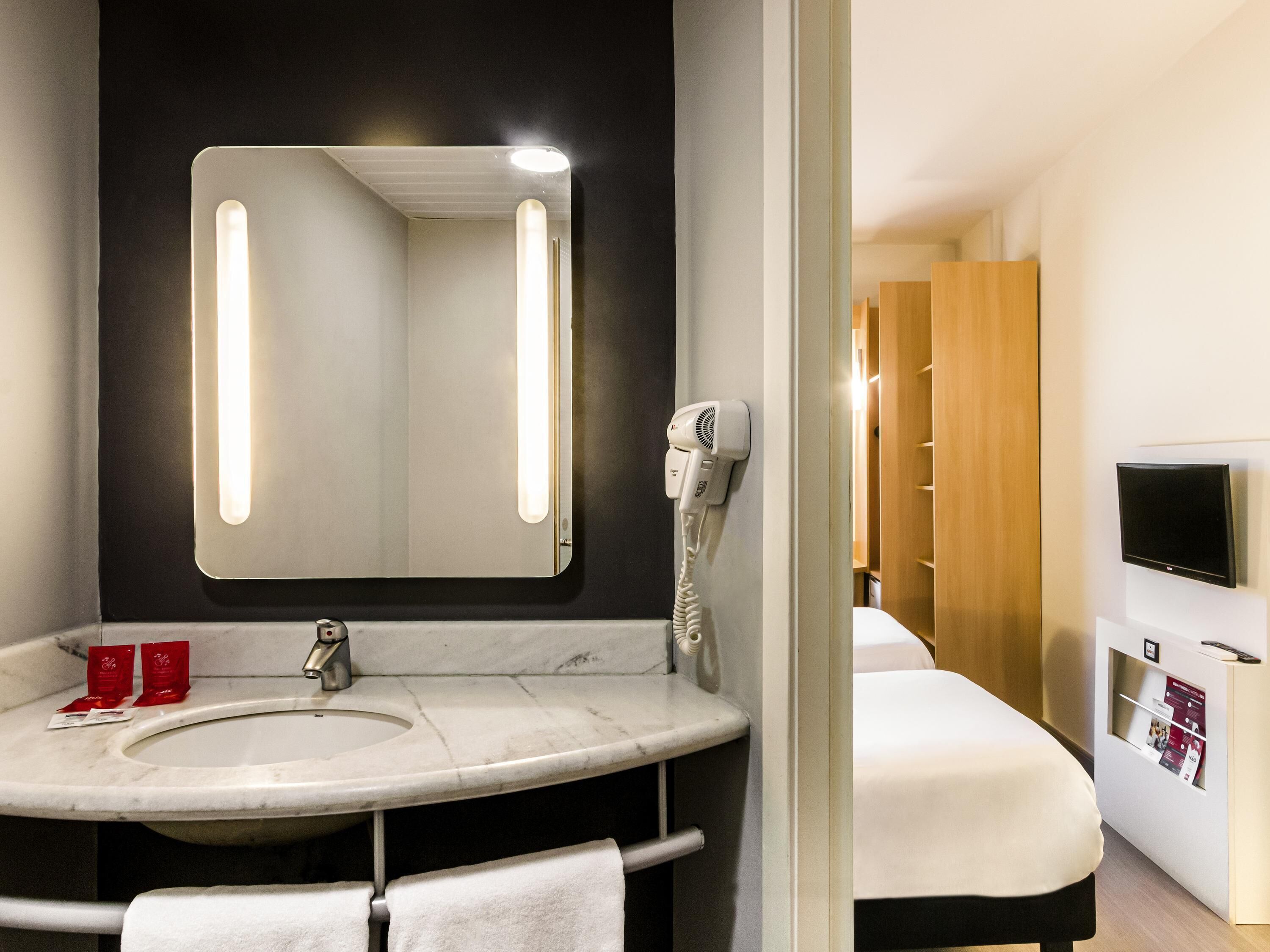 Une salle de bain d’hôtel équipée d’un lavabo, d’un miroir, d’un sèche-cheveux, et d’un lit avec un téléviseur.