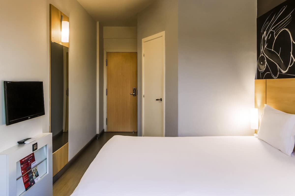 Ein Hotelzimmer mit Bett, Fernseher, Tür und Wand mit einer Leuchte.