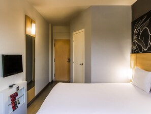 A hotel room with a bed, a TV, a door, and a wall with a light fixture.