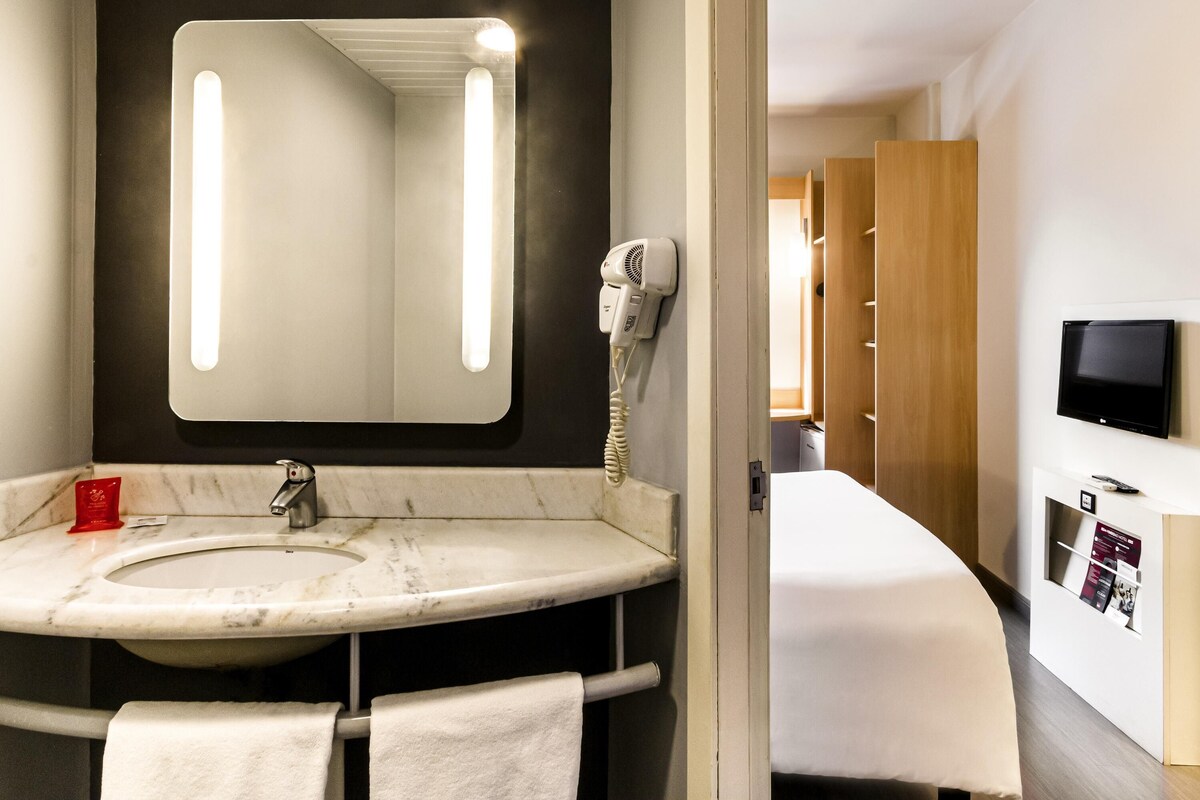 A hotel bathroom with a sink, mirror, hairdryer, and a bed with a TV.