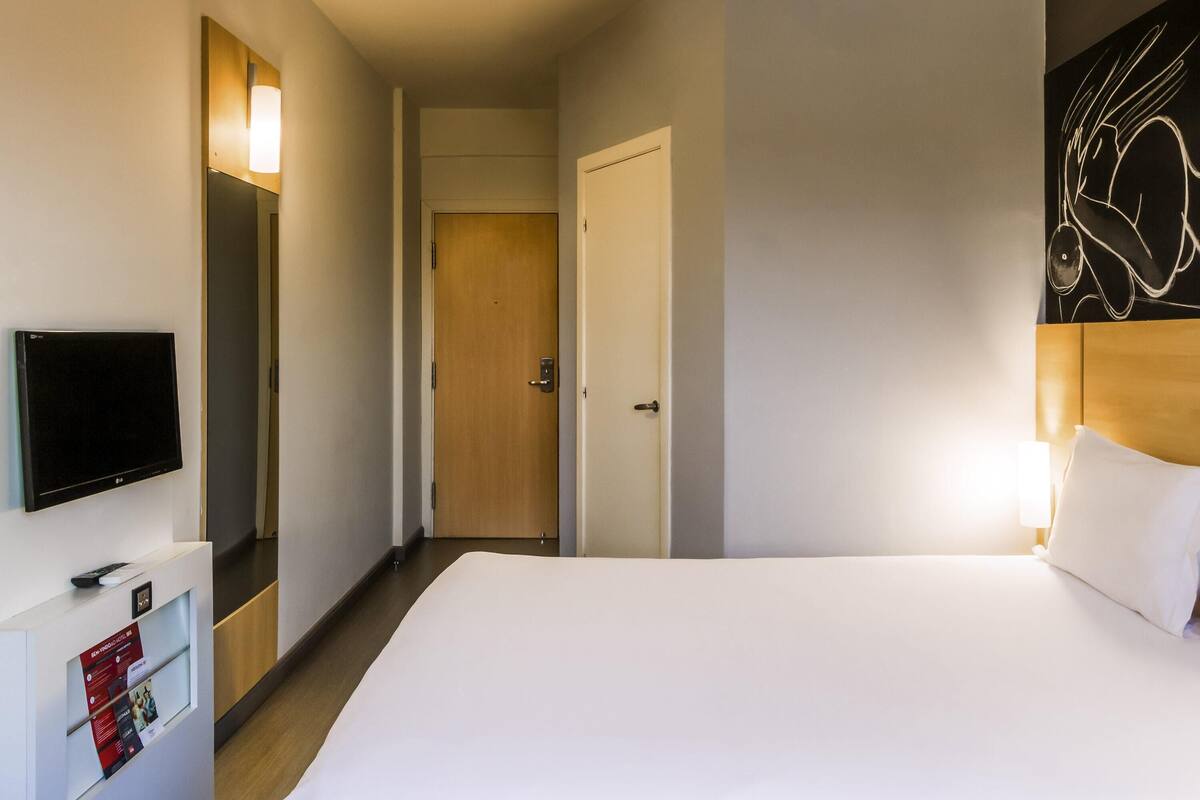 A hotel room with a bed, a TV, a door, and a wall with a light fixture.