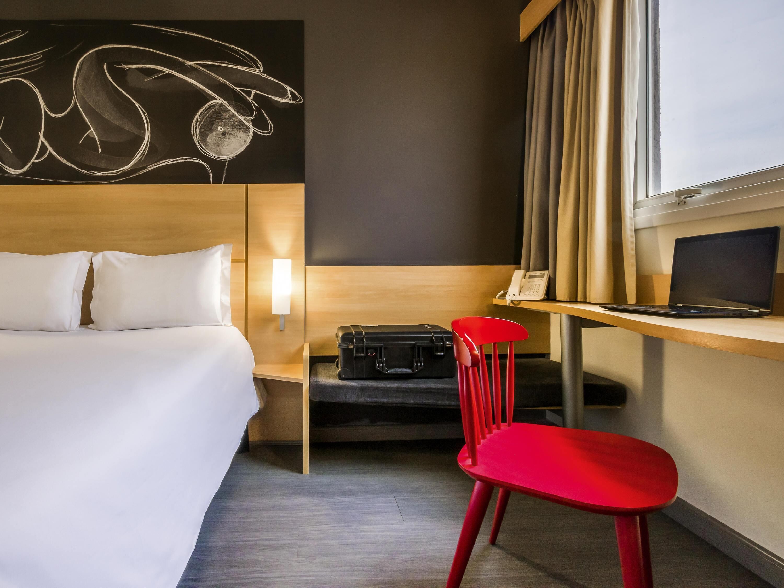 Et hotelværelse med en seng, et skrivebord med en bærbar computer, en rød stol, en kuffert og et kunstværk på væggen.