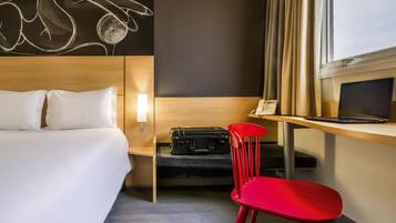 Et hotelværelse med en seng, et skrivebord med en bærbar computer, en rød stol, en kuffert og et kunstværk på væggen.