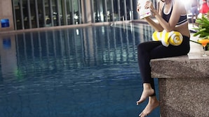 Una persona sentada junto a una piscina con una bebida, usando traje de baño y sosteniendo una toalla.