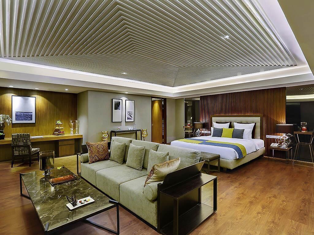 A modern hotel room with a sofa, a bed, a desk, and a ceiling with a geometric pattern.