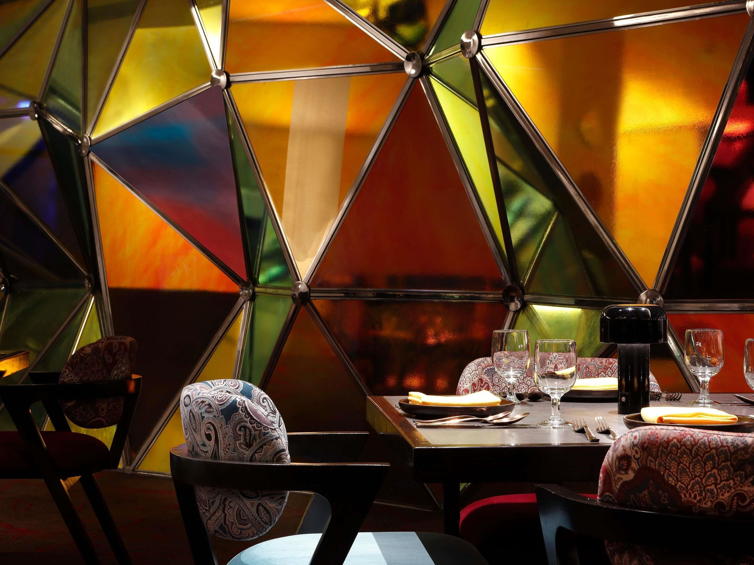 A dining area with a geometric glass partition, a table set with plates, glasses, and cutlery, and patterned chairs.