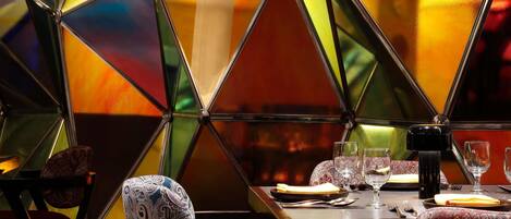 A dining area with a geometric glass partition, a table set with plates, glasses, and cutlery, and patterned chairs.