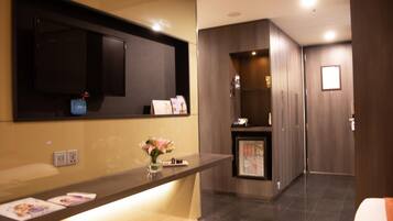 A hotel reception area with a reception desk, a small refrigerator, a mirror, and a wall-mounted television.