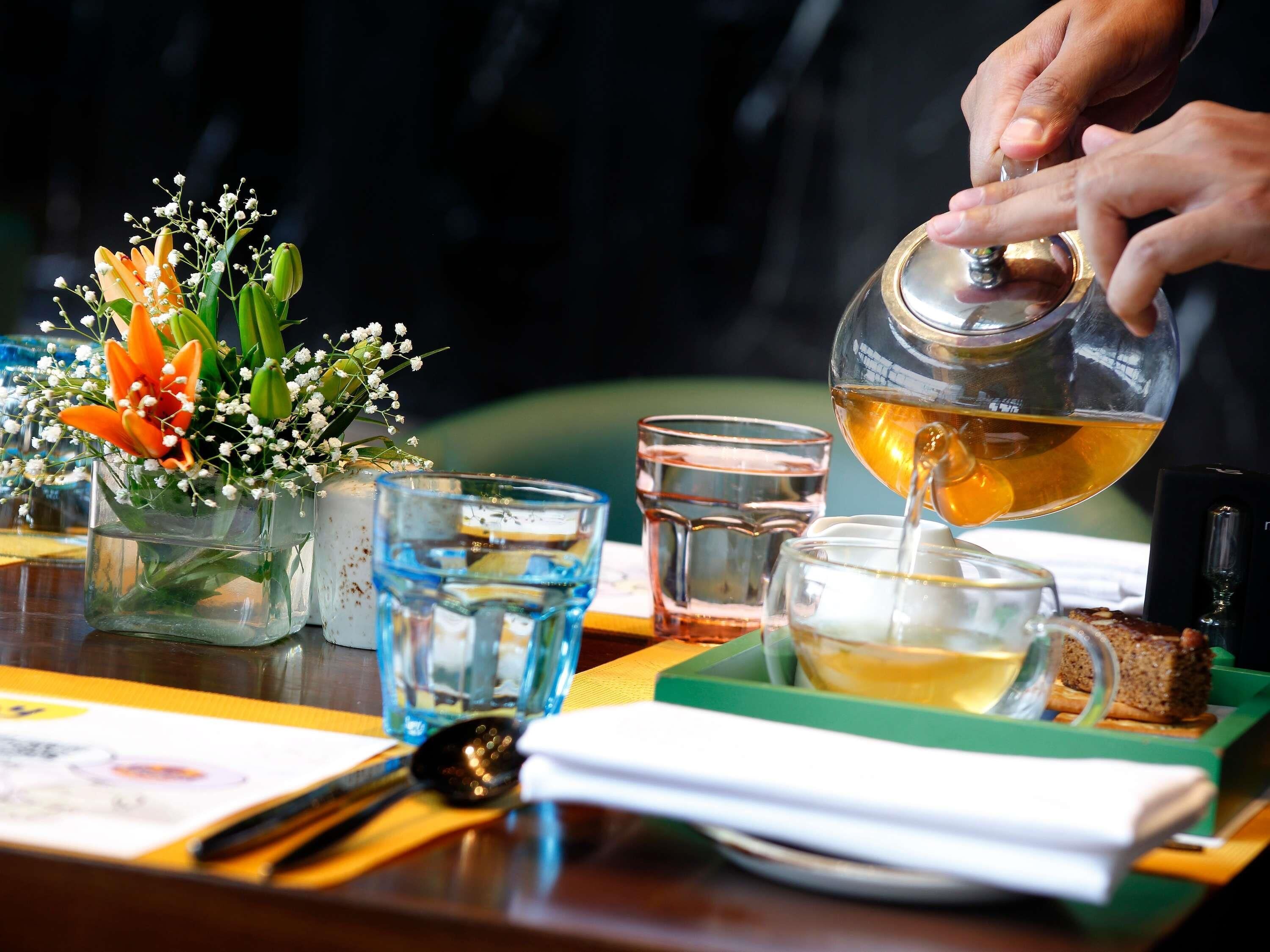 A person pouring tea into a glass, with a flower arrangement and a spoon on the table.