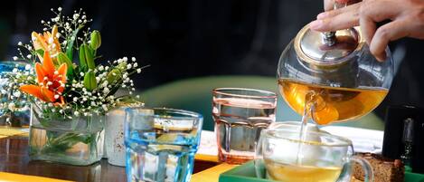A person pouring tea into a glass, with a flower arrangement and a spoon on the table.