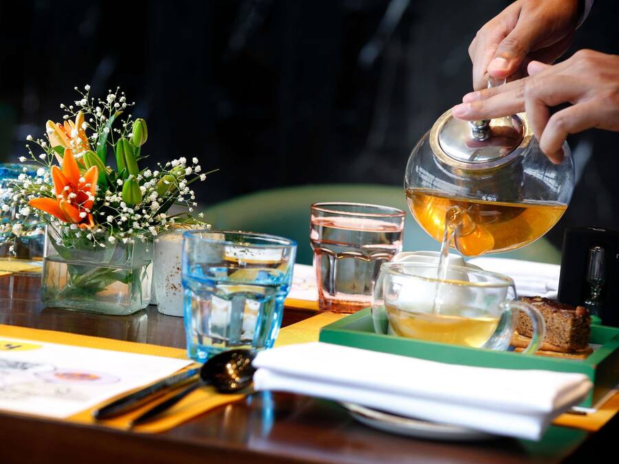 Una persona sirve té en un vaso, con un arreglo floral y una cuchara sobre la mesa.