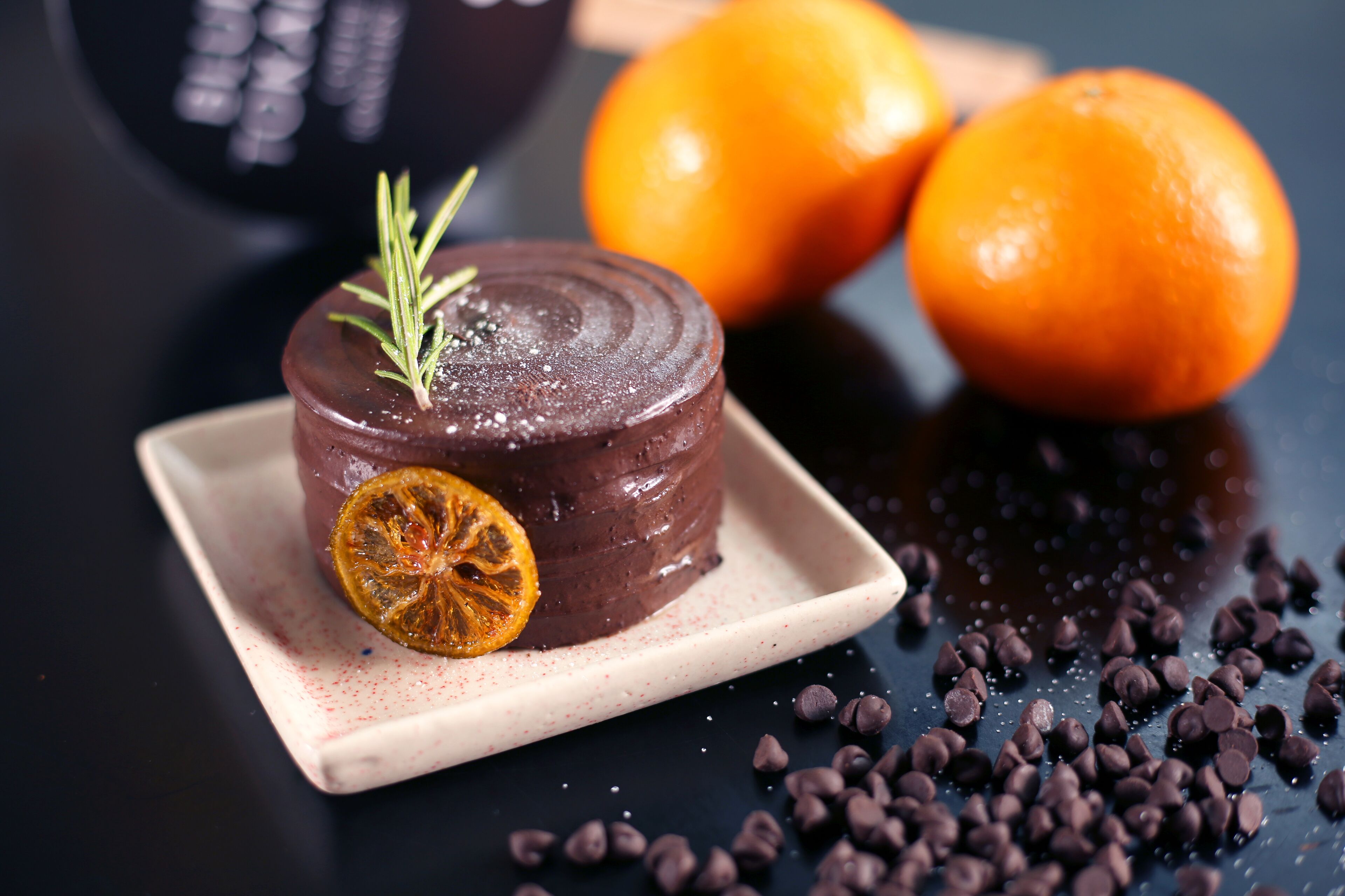 A chocolate dessert on a plate with a sprig of rosemary and a dried orange slice on top, surrounded by chocolate chips and two oranges on a dark surface.