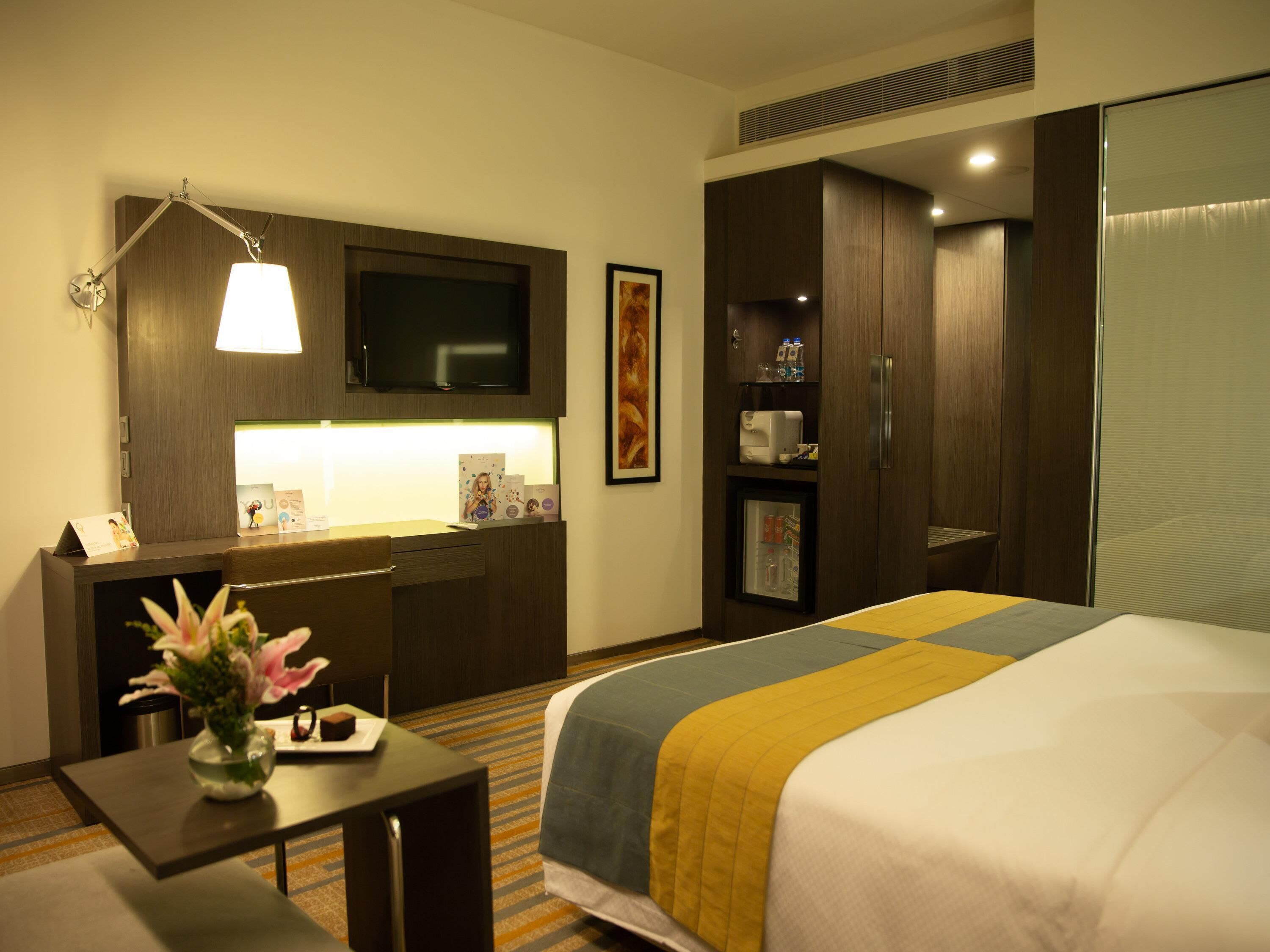 Une chambre d’hôtel équipée d’un lit, d’un bureau, d’une télévision, d’un mini-bar et d’une grande fenêtre.
