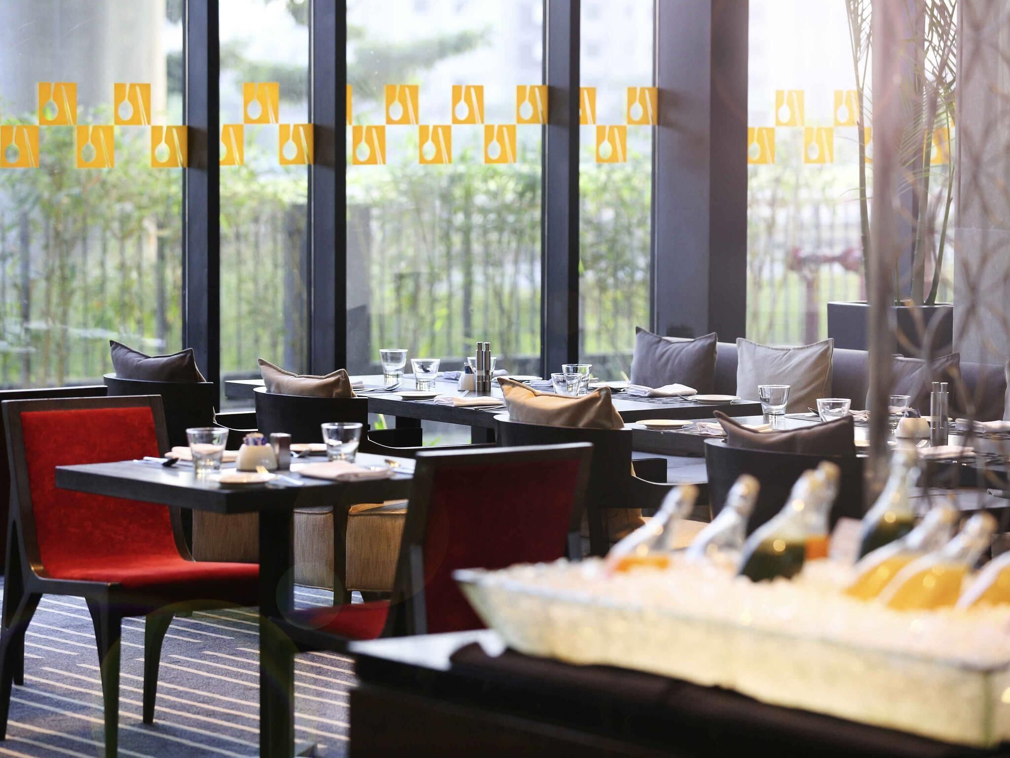 A restaurant with tables set for dining, a buffet with various dishes, and large windows with a view of greenery.