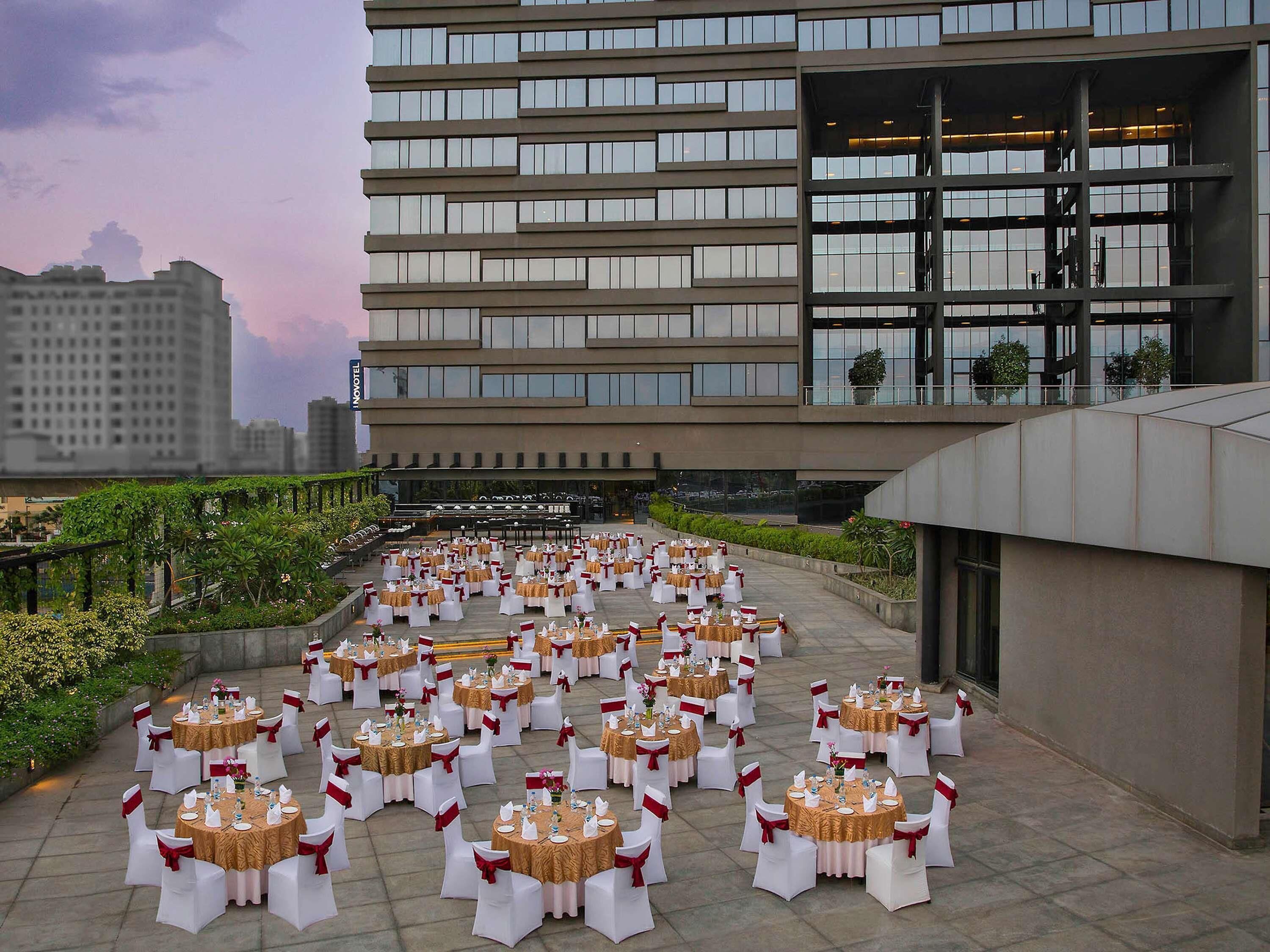 An outdoor banquet setup with round tables and white chairs, arranged on a terrace with a modern building in the background.