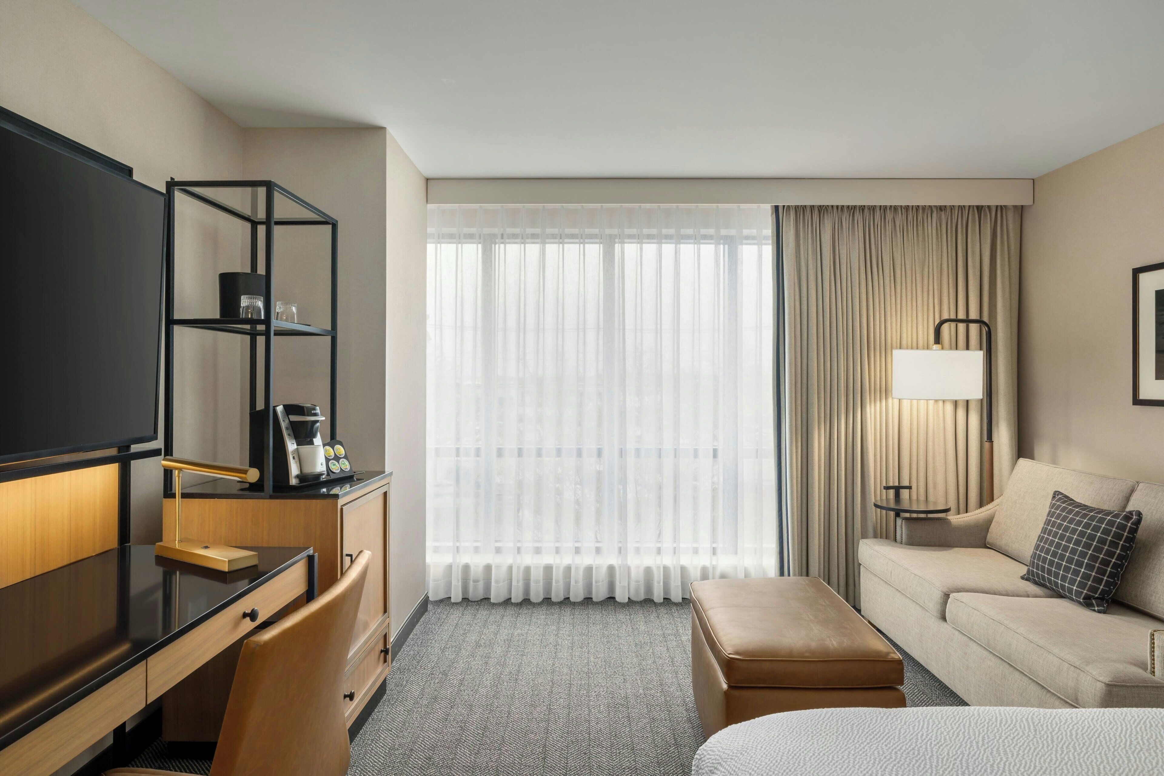 Quarto de hotel com escrivaninha, sofá, cama, televisão, máquina de café e janela com cortinas.