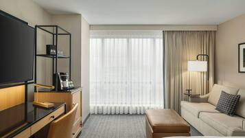 Quarto de hotel com escrivaninha, sofá, cama, televisão, máquina de café e janela com cortinas.
