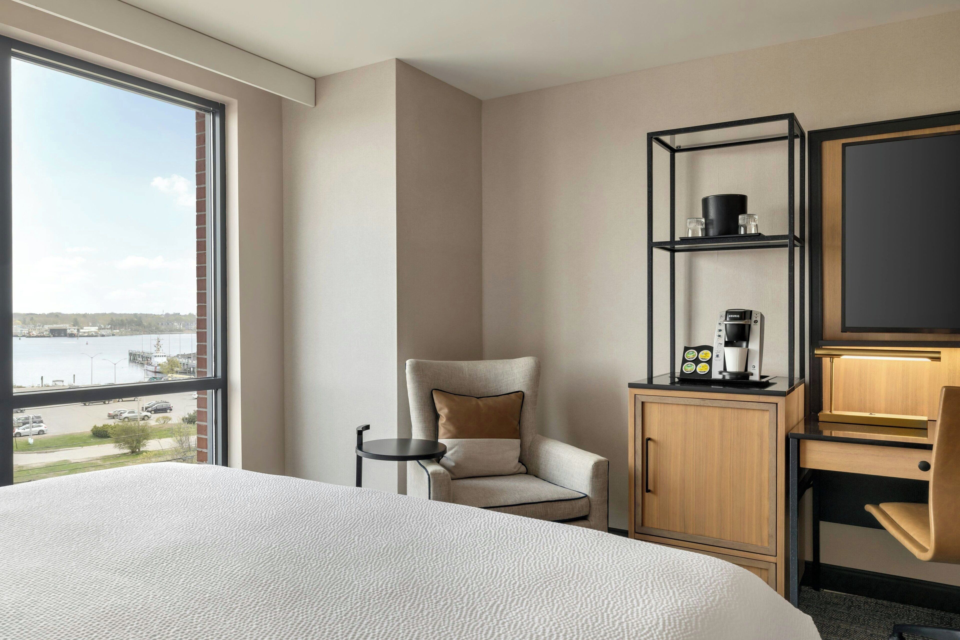 Une chambre d’hôtel avec un grand lit, une chaise, un bureau avec une machine à café, une télévision et une vue sur un plan d’eau.