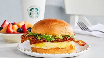 Un sándwich de desayuno en un plato, con una taza y un tazón de fruta al fondo.