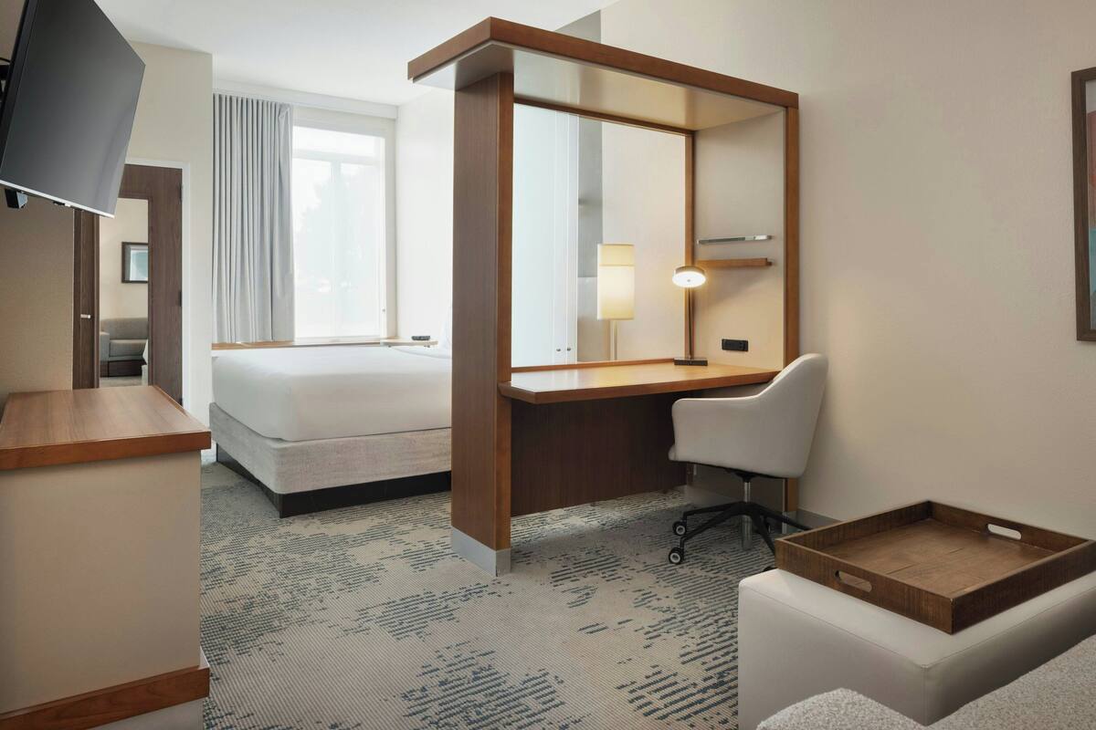 A hotel room with a bed, a desk, a chair, a TV, a mirror, and a tray table.