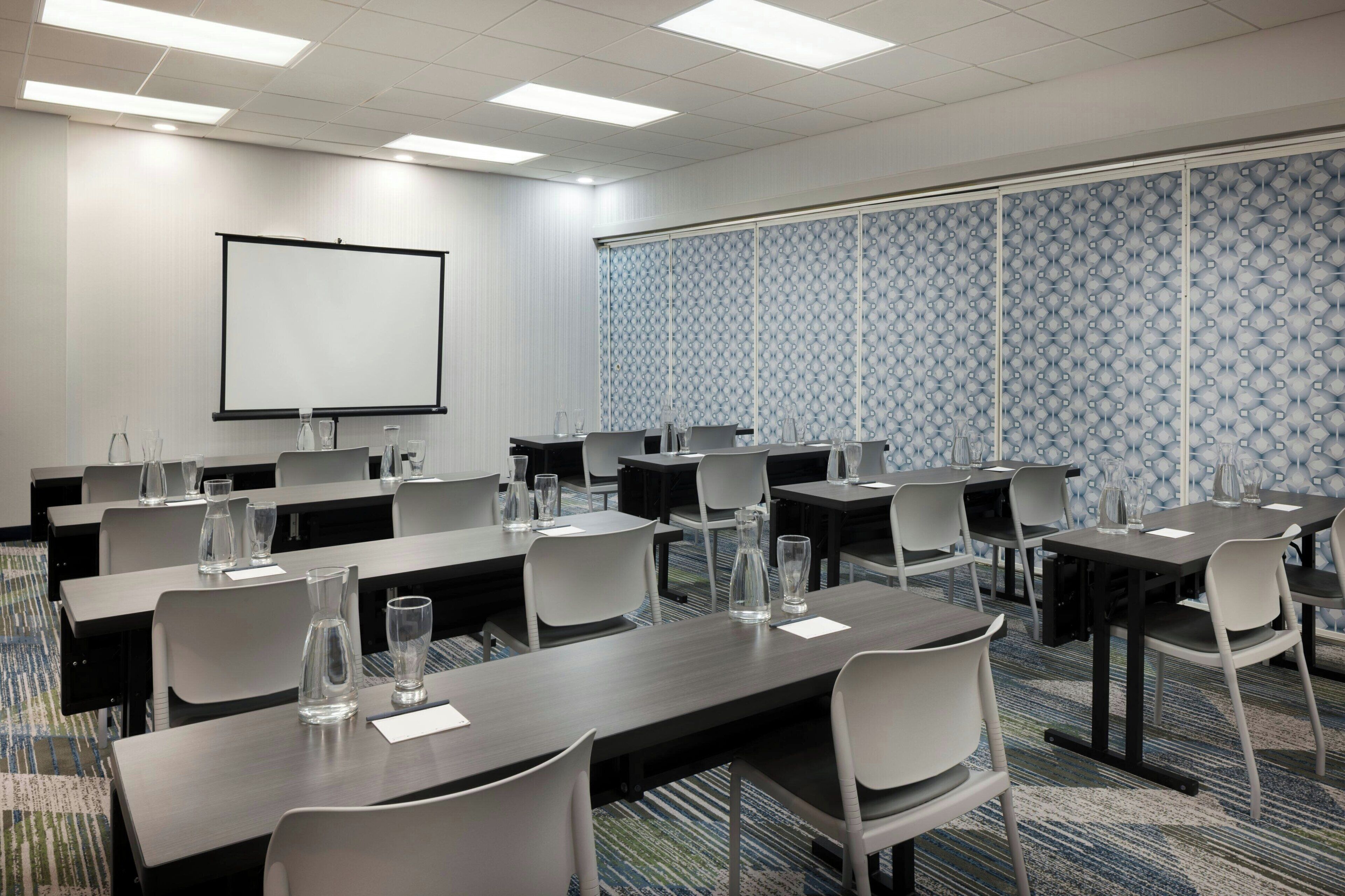 Une salle de conférence équipée de tables, de chaises, d’un écran de projection et d’un mur décoré de motifs.