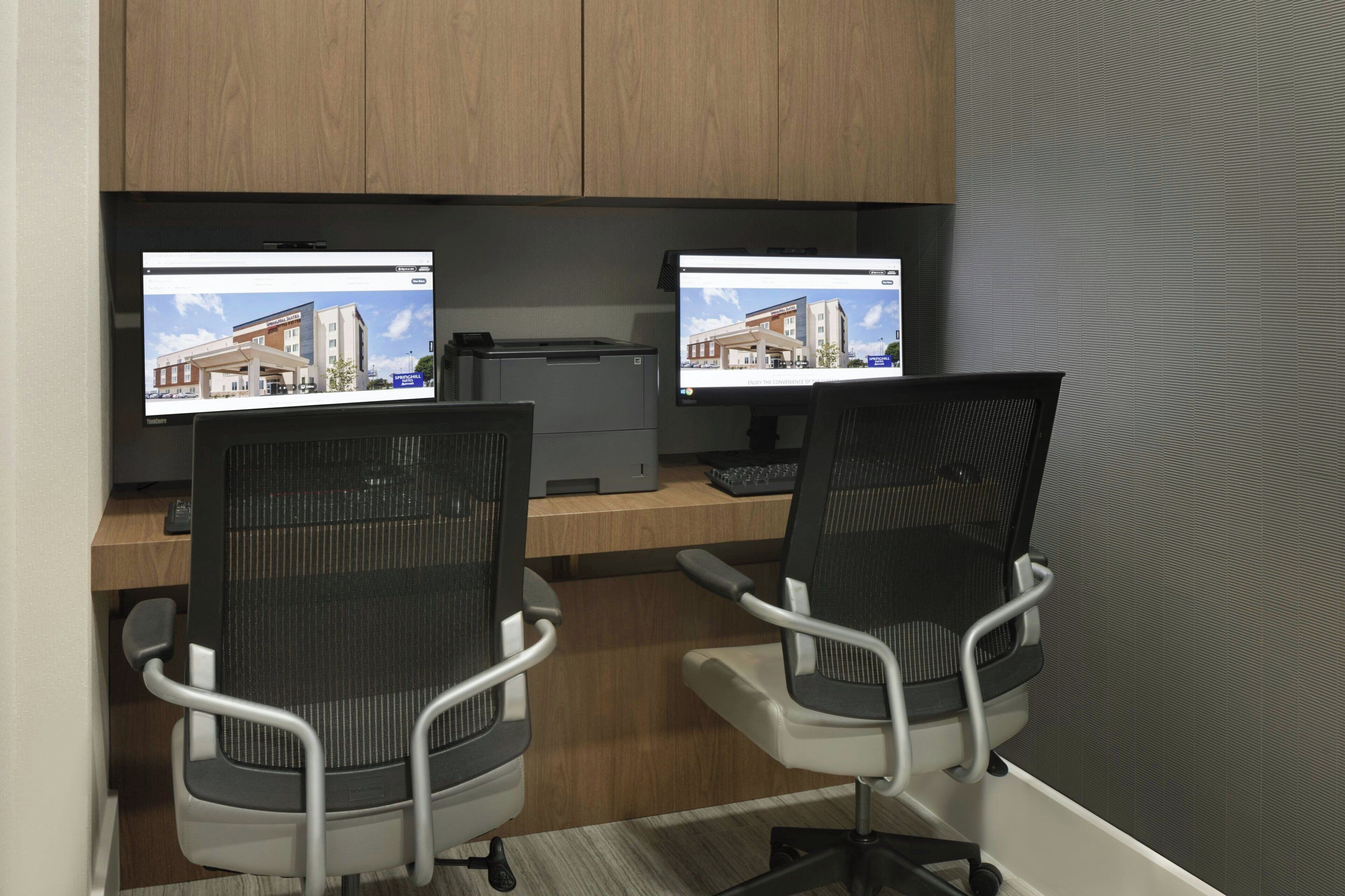 Dos sillas de oficina con respaldo de malla, un escritorio con un monitor de computadora mostrando una imagen de un edificio e impresora.