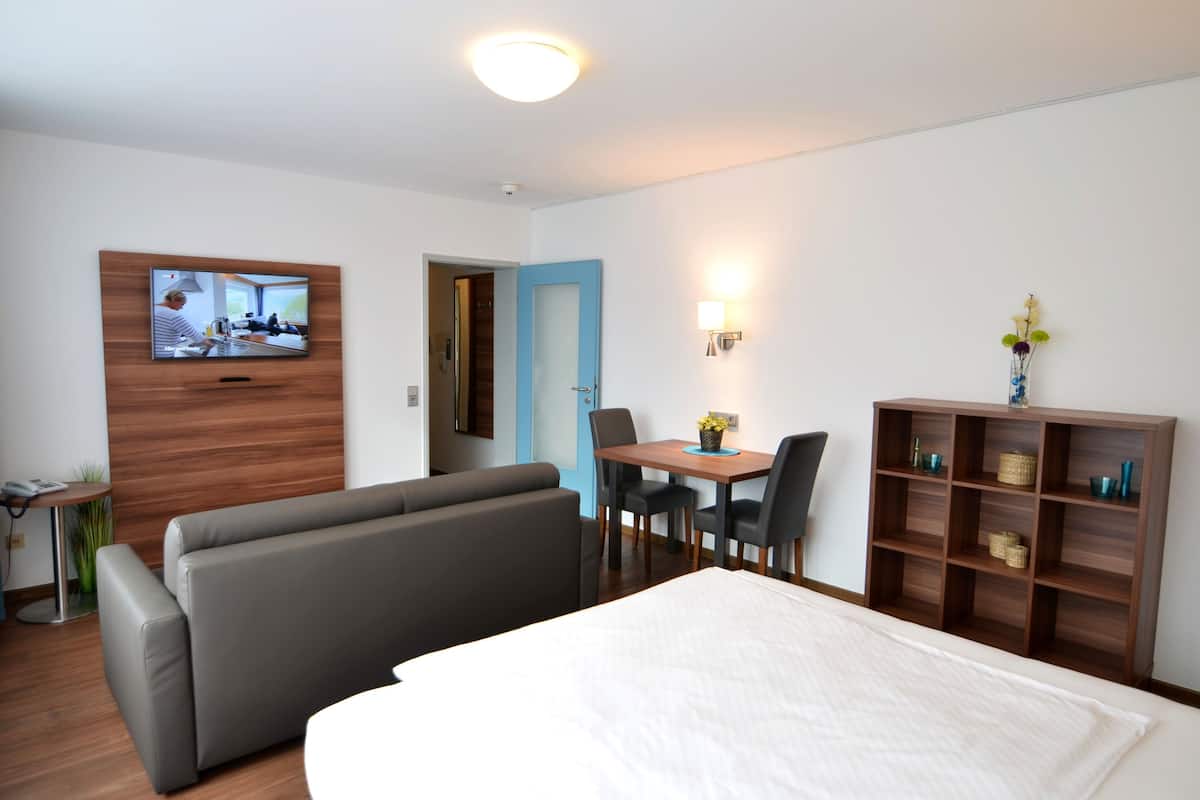Una habitación de hotel moderna con cama, sofá, mesa de comedor con sillas, televisor montado en la pared y un mueble de madera con objetos decorativos.