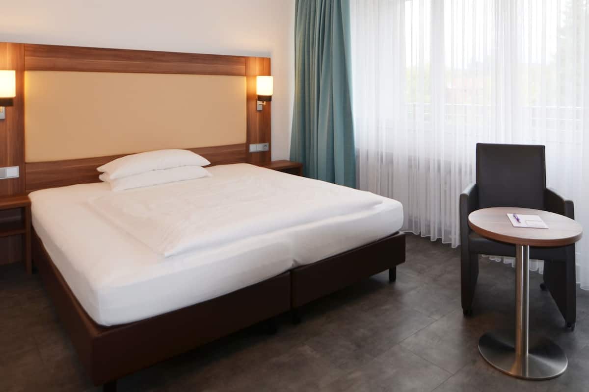 Habitación de hotel con una cama grande, dos mesitas de noche con lámparas, una pequeña mesa redonda con una silla y una ventana con cortinas traslúcidas.